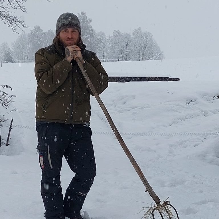 In tiefverschneiter Landschaft steht Bauer Georg lässig auf die Gabel gelehnt. Schneeflocken fallen vom Himmel während Georg zufrieden in die Kamera lächelt.
